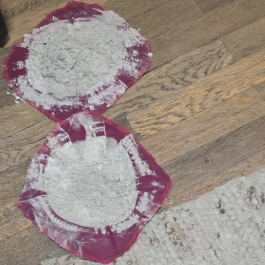 Zwei Staubfilter nach Matratzenreinigungen auf einem Holzboden, dicht mit hellem Staub bedeckt, rechts unten der Rand eines Teppichs